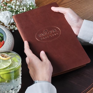 Może przedstawiać: Otwarty brązowy skórzany menu z wytłoczonym tekstem "The Parlour Dark & Oak". Na stole szklanka wody z plasterkami limonki i miska sałatki. Scena sugeruje otoczenie restauracji.
