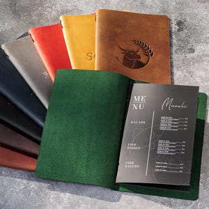 May include: A collection of leather-bound menu covers in various colors, including brown, black, red, and yellow. One cover is open, displaying a menu with the word "Menu" and the name "Muncle". The covers are arranged on a gray surface.