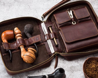 May include: A brown leather carrying case with multiple compartments for pipes and tobacco. The case is open and shows a variety of pipes and tobacco inside. There is also a small bowl of tobacco on the table.