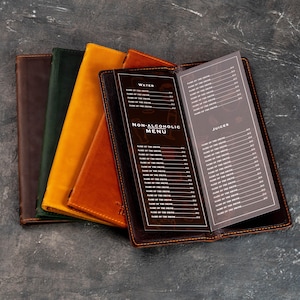 May include: A set of four leather menu covers in dark brown, forest green, mustard yellow, and tan. The open dark brown cover displays a printed non-alcoholic drink menu with sections for water, non-alcoholic drinks, and juices with listed prices.