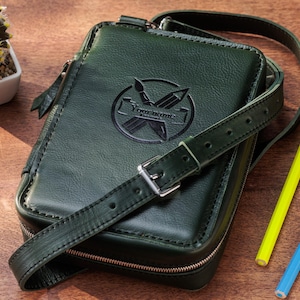May include: Dark green leather crossbody bag with a shoulder strap and a silver buckle. The bag features a debossed logo with the text "Your name" and a zipper closure. Two colored pencils, yellow and blue, are next to the bag.