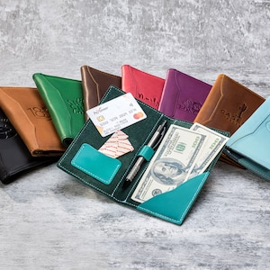 May include: A selection of leather passport holders in various colours, including black, brown, green, red, purple, and blue. The holders are arranged on a grey textured surface. One holder is open, displaying a credit card, pen, and cash.