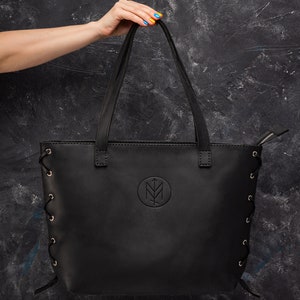 May include: Black leather tote bag with a logo and lace-up sides. The bag has a zipper closure and two top handles.