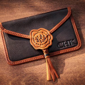 May include: Black leather wallet with brown trim and a decorative seal. The wallet features a tassel and the initials "CHK". The design is set against a wooden background.
