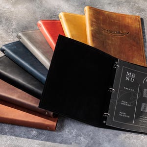 May include: A collection of leather menu covers in various colors including black, brown, tan, red, gray, and blue. One black cover is open, revealing a three-ring binder mechanism holding a transparent sheet with the text MENU and Muesli.