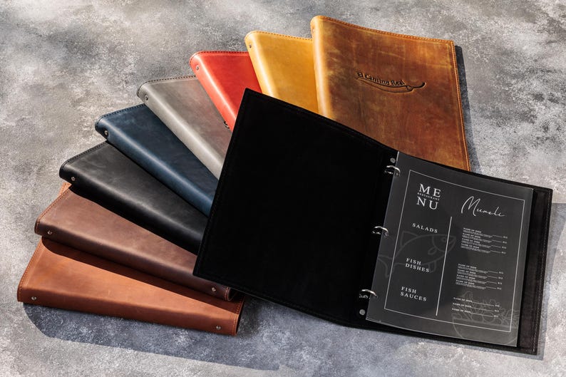 May include: A collection of leather menu covers in various colors including black, brown, tan, red, gray, and blue. One black cover is open, revealing a three-ring binder mechanism holding a transparent sheet with the text MENU and Muesli.