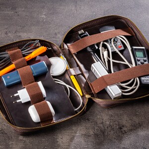 May include: A brown leather zipped case with multiple compartments and elastic straps for organising electronics and accessories. The case is open and filled with various items, including a black and silver power bank, a white charging cable, a black remote control, and a silver USB adapter.