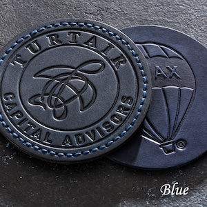 May include: Two round leather coasters, one with a blue stitched border and the text "TURTAIR CAPITAL ADVISORS" and a stylized turtle design, the other with a hot air balloon design and the text "AX" in the basket.