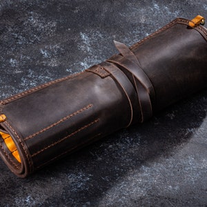 K&ouml;nnte beinhalten: Eine braune Lederrolle mit gelben N&auml;hten und einem Messing-Schnappverschluss. Die Rolle ist aufgerollt und mit einem Lederriemen gebunden.
