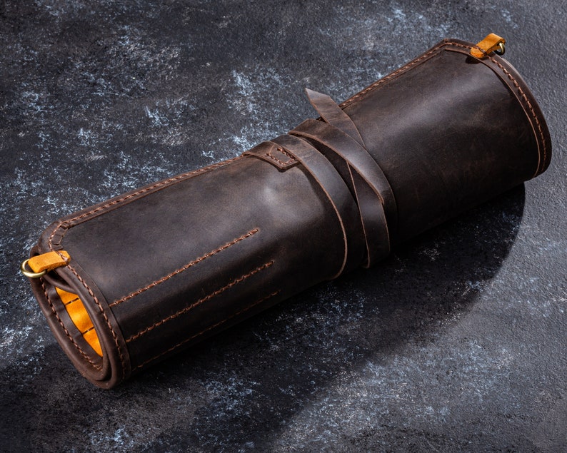 K&ouml;nnte beinhalten: Eine braune Lederrolle mit gelben N&auml;hten und einem Messing-Schnappverschluss. Die Rolle ist aufgerollt und mit einem Lederriemen gebunden.