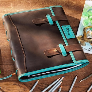 May include: A brown leather journal with teal accents, buckles, and the name "Maria" on a teal patch. A sketchbook with a cat illustration and colored pencils are on a wooden surface.