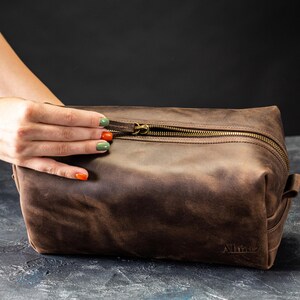 May include: A brown leather toiletry bag with a brass zipper and a small handle on the side. The bag has a rectangular shape and is sitting on a dark surface. The word "Allianz" is embossed on the bottom.