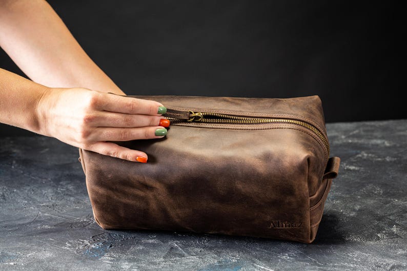 May include: A brown leather toiletry bag with a brass zipper and a small handle on the side. The bag has a rectangular shape and is sitting on a dark surface. The word "Allianz" is embossed on the bottom.