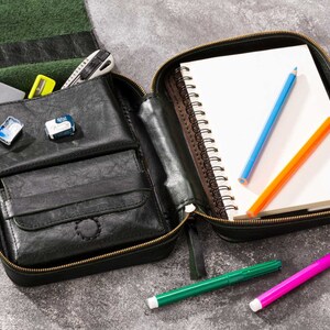 May include: A black leather zippered pouch with a notebook, pencils, and markers inside. The notebook has a white cover and a spiral binding. The pencils are blue, orange, and green. The markers are green and pink.