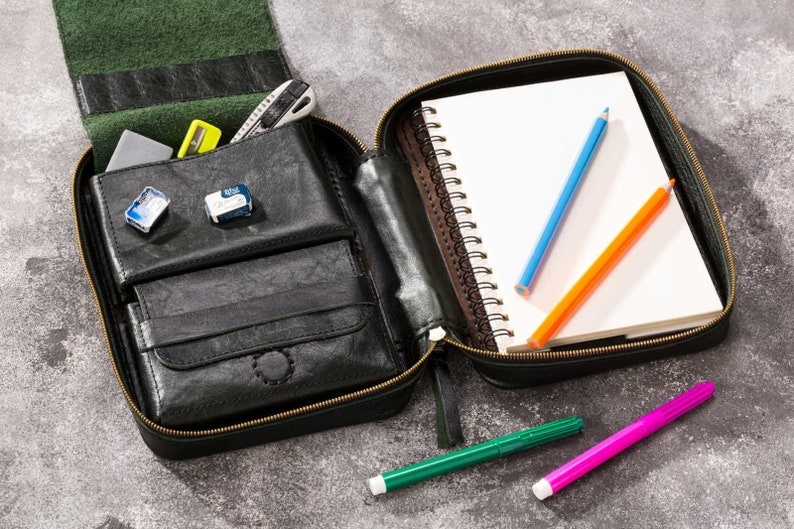 May include: A black leather zippered pouch with a notebook, pencils, and markers inside. The notebook has a white cover and a spiral binding. The pencils are blue, orange, and green. The markers are green and pink.