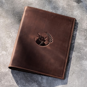 May include: A dark brown leather folder featuring a debossed circular emblem on the front cover. The emblem depicts the head of a bull positioned next to a stalk of wheat. The folder has visible stitching along the edges and a smooth finish.