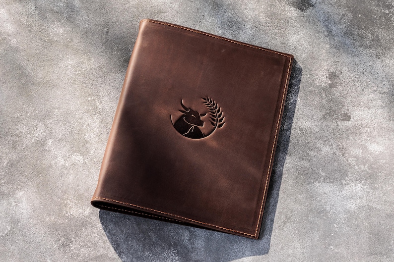 May include: A dark brown leather folder featuring a debossed circular emblem on the front cover. The emblem depicts the head of a bull positioned next to a stalk of wheat. The folder has visible stitching along the edges and a smooth finish.