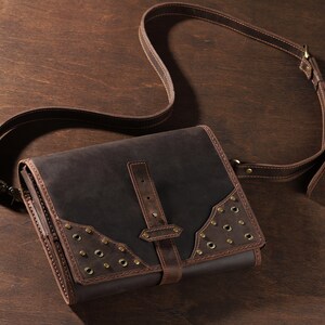May include: A dark brown leather satchel with a long adjustable strap. The bag features a flap closure with a buckle and decorative metal rivets. The bag is made of a durable leather material and is designed for carrying items.