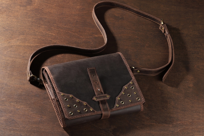 May include: A dark brown leather satchel with a long adjustable strap. The bag features a flap closure with a buckle and decorative metal rivets. The bag is made of a durable leather material and is designed for carrying items.