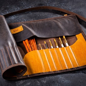 K&ouml;nnte beinhalten: Eine braune Lederrolle mit Klappe und Riemen. Die Rolle hat ein personalisiertes Etikett mit der Aufschrift "BJM" und ist mit Pinseln gef&uuml;llt.