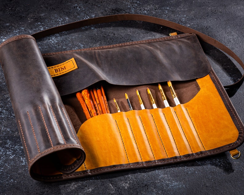 K&ouml;nnte beinhalten: Eine braune Lederrolle mit Klappe und Riemen. Die Rolle hat ein personalisiertes Etikett mit der Aufschrift "BJM" und ist mit Pinseln gef&uuml;llt.