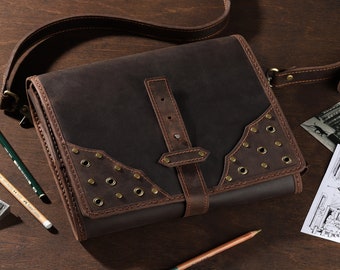 May include: A dark brown leather messenger bag with a shoulder strap, buckle closure, and brass rivets. The bag is rectangular with decorative stitching and is on a wooden surface. Pencils and a pencil sharpener are also present.