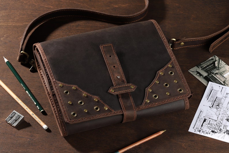 May include: A dark brown leather messenger bag with a shoulder strap, buckle closure, and brass rivets. The bag is rectangular with decorative stitching and is on a wooden surface. Pencils and a pencil sharpener are also present.