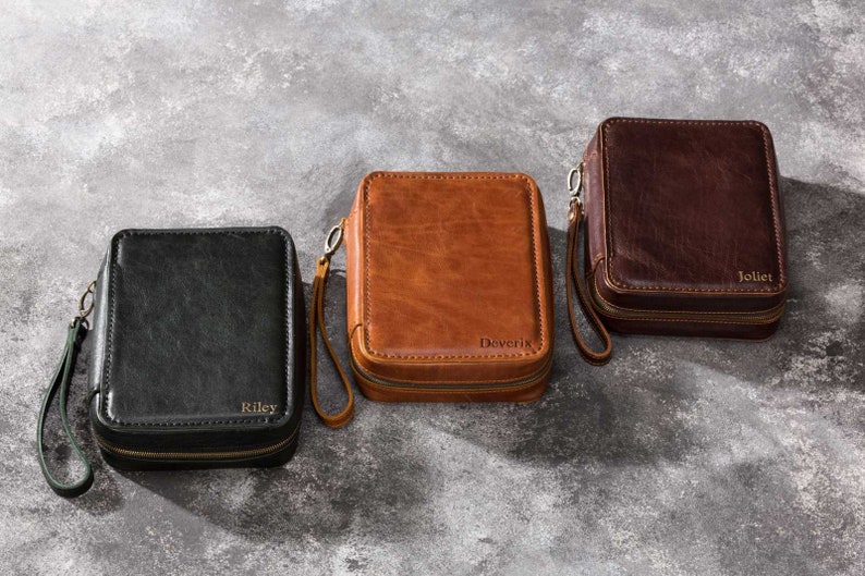 May include: Three personalised leather zippered pouches in black, brown, and dark brown. The pouches have a zipper closure and a small handle. The pouches are labelled with the names "Riley", "Doverix", and "Jolliet".