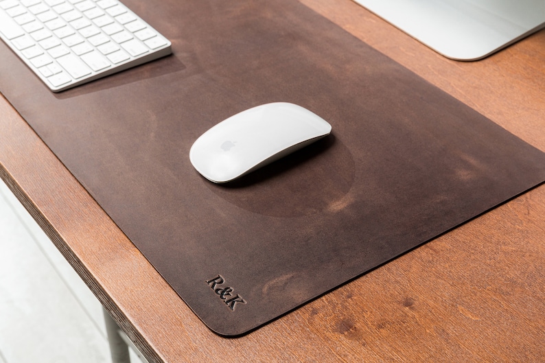 May include: A brown leather desk mat with the text "R&K" embossed on the bottom right corner. A white wireless mouse is on the mat.