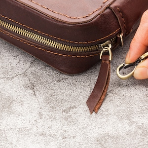 May include: A brown leather zipped pouch with a brass zipper pull and a leather strap with a brass hook.
