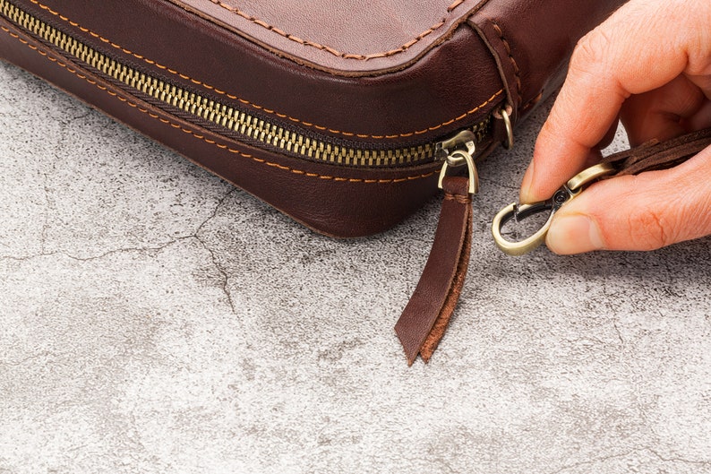 May include: A brown leather zipped pouch with a brass zipper pull and a leather strap with a brass hook.