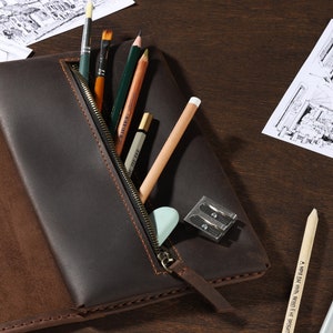 May include: A dark brown leather pencil case with a zipper, open to reveal art supplies. The case holds pencils, paintbrushes, an eraser, and a pencil sharpener. Architectural sketches are visible in the background.