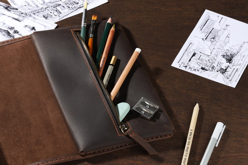 May include: A dark brown leather pencil case with a zipper, open to reveal art supplies. The case holds pencils, paintbrushes, an eraser, and a pencil sharpener. Architectural sketches are visible in the background.