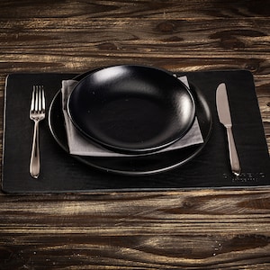 May include: A black placemat with a black plate and a silver fork and knife. The place setting is on a wooden table.