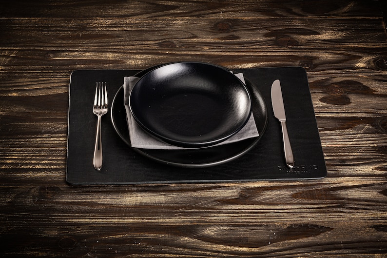 May include: A black placemat with a black plate and a silver fork and knife. The place setting is on a wooden table.