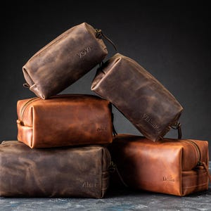 May include: A stack of personalized leather toiletry bags in various shades of brown. Each bag has a zipper closure and a small handle. The names "Yova", "Alex", "Vladimir", "Almer", and "Denis" are embossed on the bags.