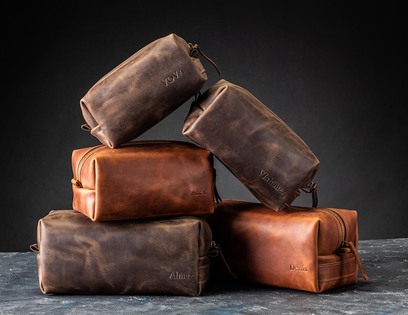May include: A collection of five leather toiletry bags in various shades of brown. Each bag has a zipper closure and a small handle. The bags are personalized with names like "Yova", "Alex", "Vladimir", "Almer", and "Denis" embossed on them.