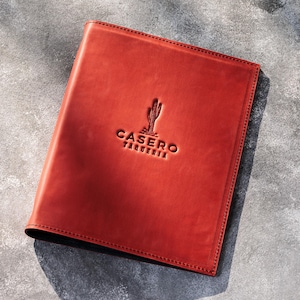 May include: A rectangular, reddish-brown leather menu cover featuring a debossed cactus icon and the text CASERO TAQUERIA centered on the front. The cover has visible stitching along the edges and a smooth, matte finish on the surface.