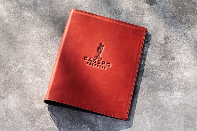 May include: A rectangular, reddish-brown leather menu cover featuring a debossed cactus icon and the text CASERO TAQUERIA centered on the front. The cover has visible stitching along the edges and a smooth, matte finish on the surface.