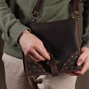 May include: A dark brown leather messenger bag with a shoulder strap. The bag features a flap closure with a buckle and decorative brass rivets. The bag is held by a person wearing a green sweater and khaki pants.