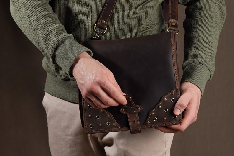 May include: A dark brown leather messenger bag with a shoulder strap. The bag features a flap closure with a buckle and decorative brass rivets. The bag is held by a person wearing a green sweater and khaki pants.