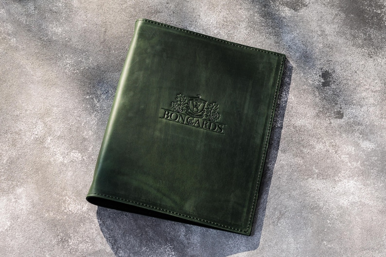 May include: A dark forest green leather folder featuring a rectangular shape with stitched edges. The front cover displays a debossed emblem with the text BONGARDS centered in the middle, surrounded by a crest design with leaf-like details.