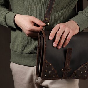 May include: A dark brown leather messenger bag with a flap closure and a shoulder strap. The bag features decorative rivets and brown leather accents. The person is wearing a green sweater and khaki pants.