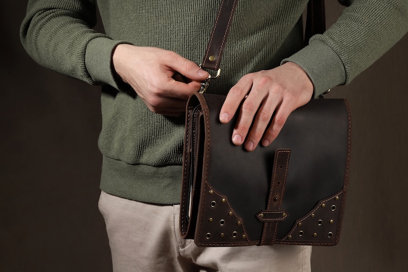 May include: A dark brown leather messenger bag with a flap closure and a shoulder strap. The bag features decorative rivets and brown leather accents. The person is wearing a green sweater and khaki pants.