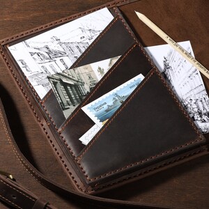 May include: A dark brown leather notebook with multiple pockets, holding postcards, stamps, and a pencil. The notebook is open, revealing the contents. The pencil has text on it. The notebook is on a wooden surface.
