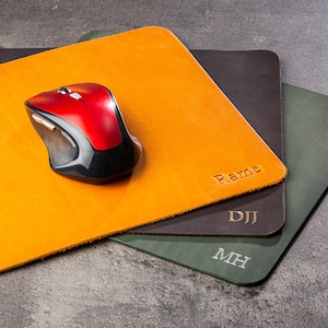 May include: A red and black wireless computer mouse rests on a square, orange leather mousepad. Two additional leather mousepads in dark brown and green are visible. Each pad has initials engraved.
