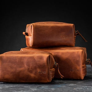 May include: Three stacked, rectangular, brown leather toiletry bags. The bags have a zipper closure and a small handle. The names "Alex", "Denis", and "Nastyc" are embossed on the bags. The bags are on a dark surface.