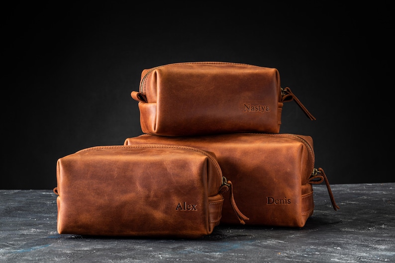 May include: Three stacked, rectangular, brown leather toiletry bags. The bags have a zipper closure and a small handle. The names "Alex", "Denis", and "Nastyc" are embossed on the bags. The bags are on a dark surface.