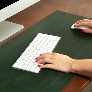 May include: A dark green desk mat with a white keyboard and mouse. The mat is rectangular and covers a portion of a wooden desk. The keyboard is in the center, and the mouse is to the right. The computer monitor is in the background.