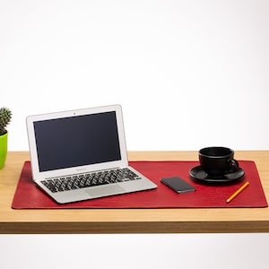 May include: A modern office desk setup featuring a laptop, smartphone, and a black coffee cup on a red leather desk mat. A small cactus in a green pot and a yellow pencil complete the workspace. The desk is made of light wood.